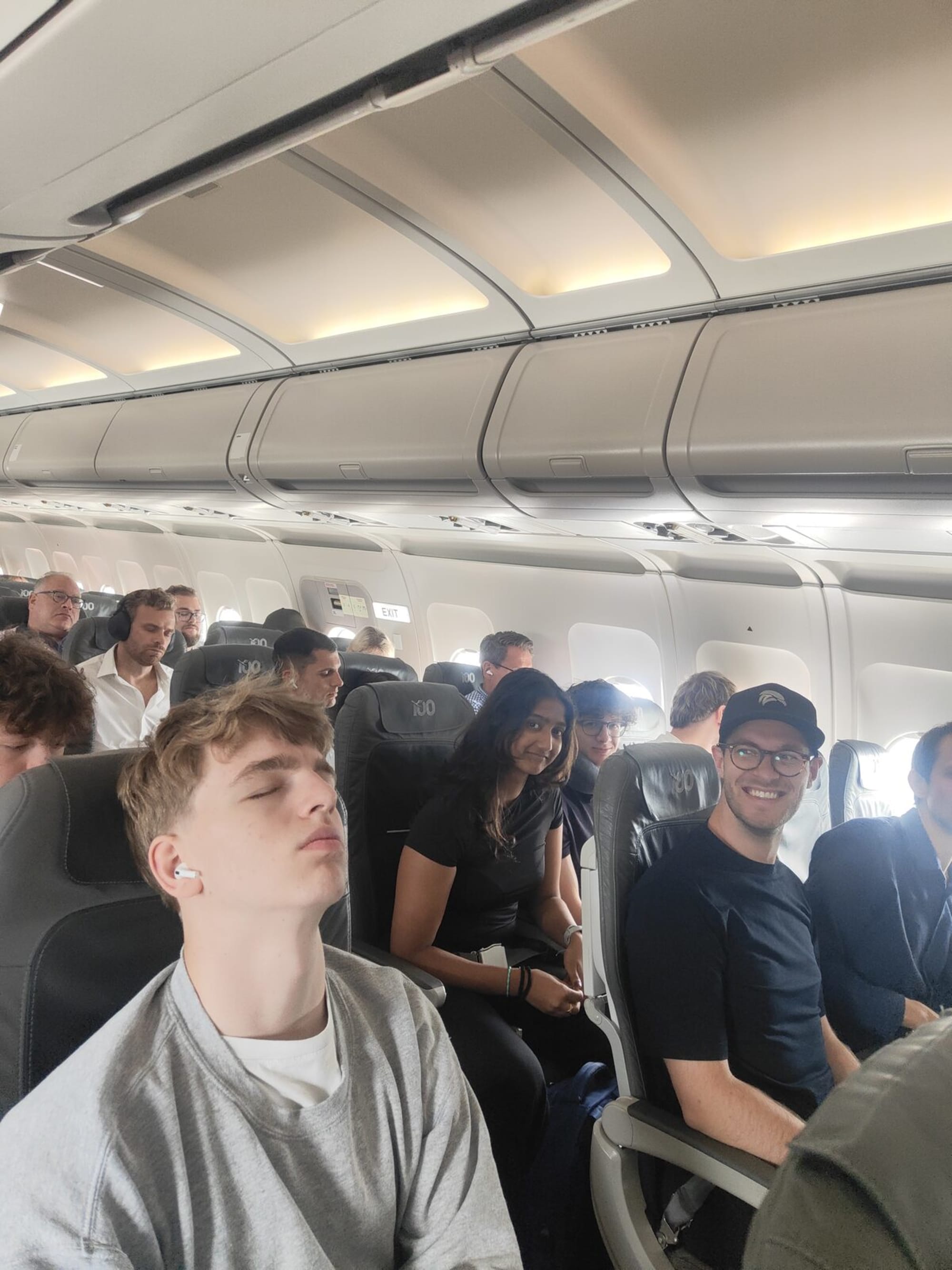 Teilnehmer:innen in der Flugzeugkabine — einer schläft, einer lacht in die Kamera